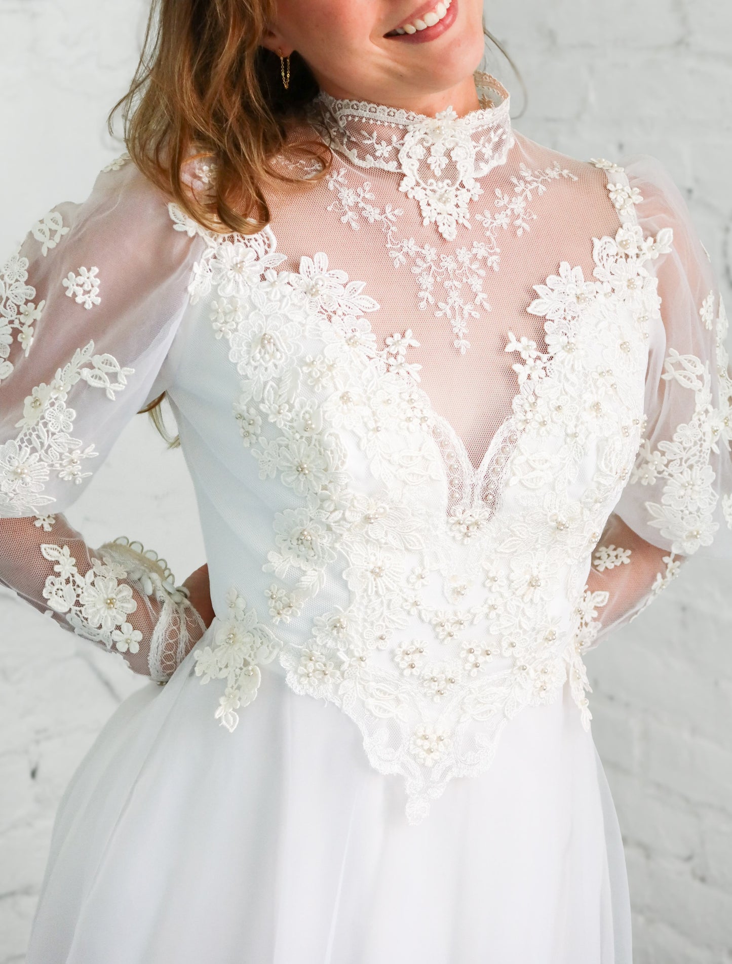 A close-up of a woman wearing a white lace and tulle wedding dress with a high mock neck, sheer long sleeves, and a fitted bodice with floral lace applique.
