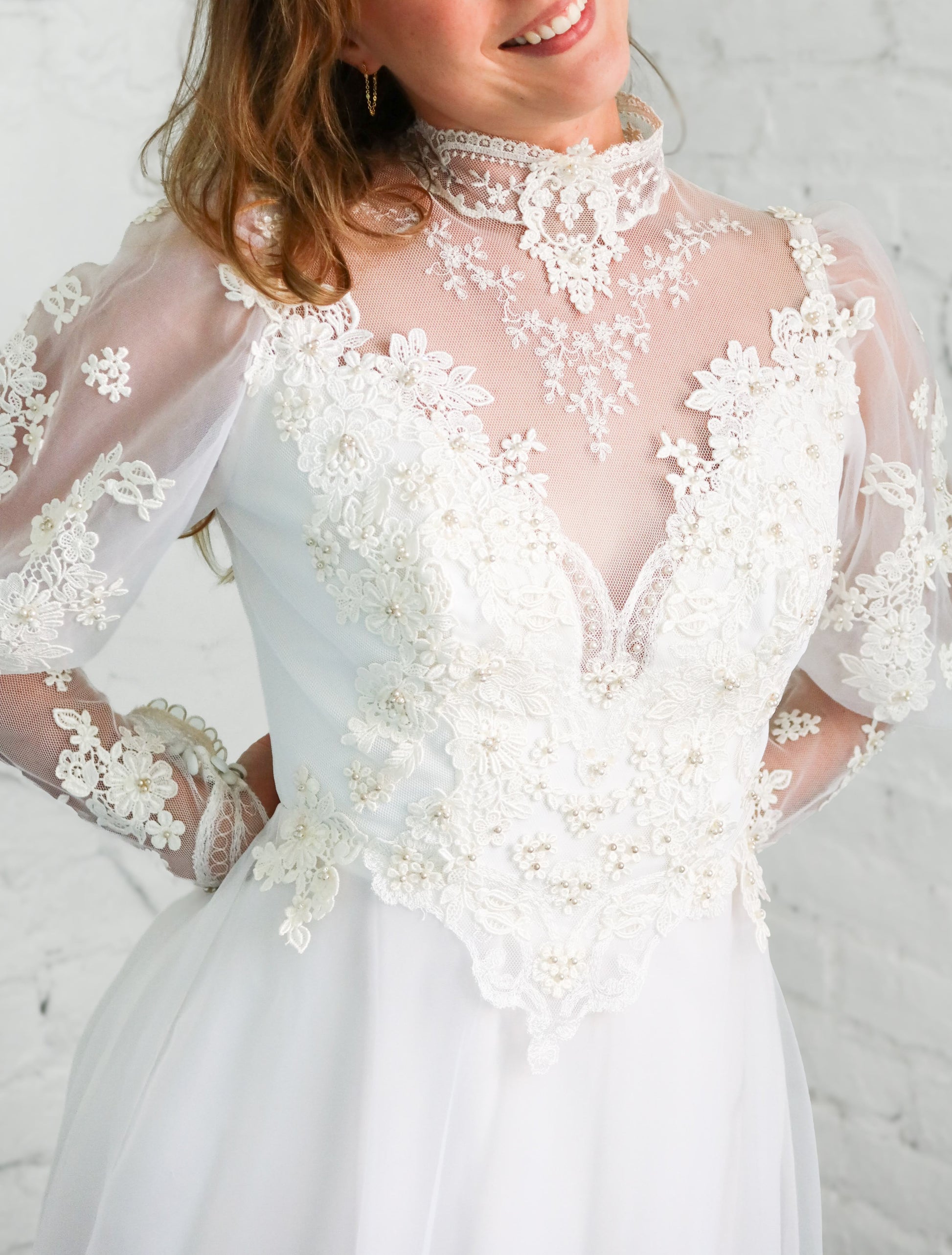 A close-up of a woman wearing a white lace and tulle wedding dress with a high mock neck, sheer long sleeves, and a fitted bodice with floral lace applique.